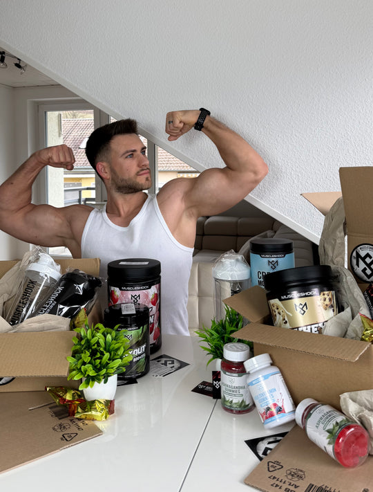 Ein Mann posiert mit angespannten Oberarmen hinter Muscleshock Supplements und geöffneten Versandkartons.