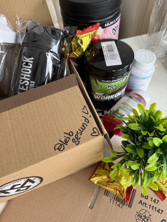 Geöffneter Muscleshock Versandkarton mit Shaker, umgeben von Supplements; Aufschrift: Bleib gesund.
