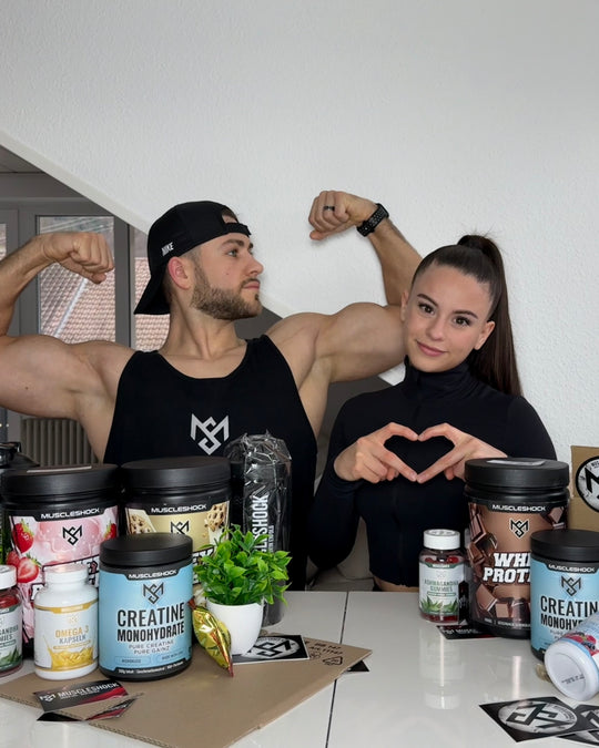 Ein Mann zeigt seine Oberarme, eine Frau formt ein Herz mit den Händen; Muscleshock Supplements auf dem Tisch.