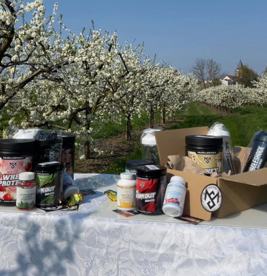 Muscleshock Supplements und ein geöffneter Versandkarton auf einem Tisch vor blühenden Obstbäumen.