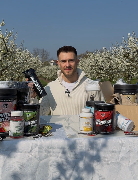 Ein Mann zeigt einen schwarzen Muscleshock Shaker an einem Tisch mit Supplements vor blühenden Bäumen.